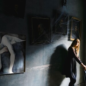 May include: A dark room features a large rectangular painting of a reclining figure with pale skin and flowing hair. Three framed portraits hang on the wall above, partially covered in artificial spider webs. A person stands nearby.