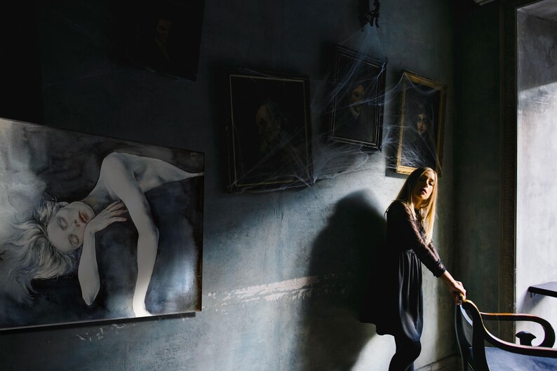 May include: A dark room features a large rectangular painting of a reclining figure with pale skin and flowing hair. Three framed portraits hang on the wall above, partially covered in artificial spider webs. A person stands nearby.