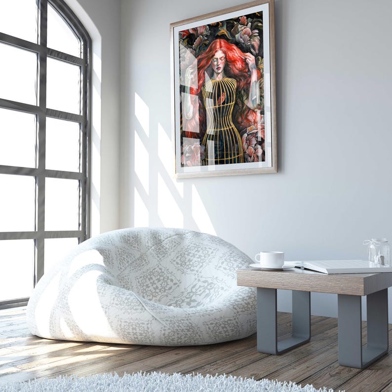 Op de afbeelding: Een ingelijste muurkunstprint met een persoon met lang rood haar in een gouden vogelkooi, omringd door donkere florale elementen. Het kunstwerk is ingelijst in een lichthouten lijst aan een witte muur boven een witte zitzak met patroon.