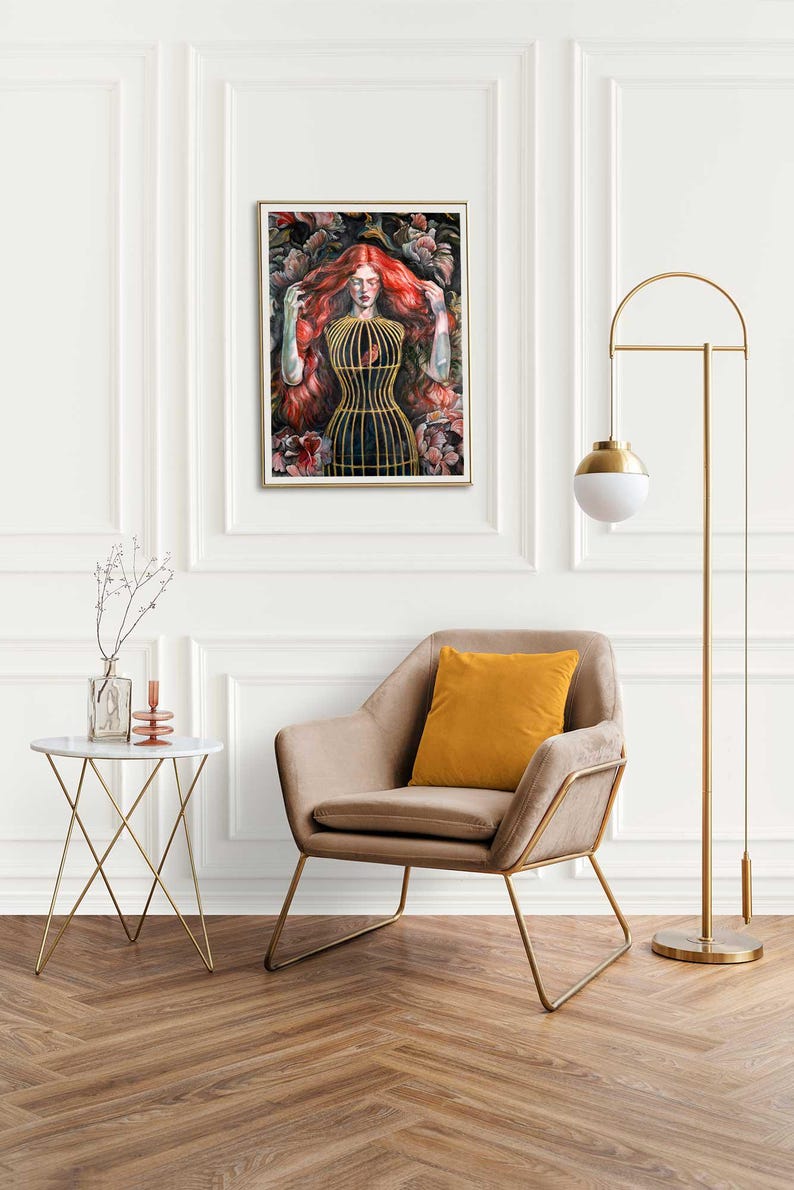 Op de afbeelding: Een ingelijste kunstdruk met een schilderij van een persoon met lang rood haar die een goudkleurig kooivormig lijfje draagt, omringd door donkere florale elementen. Het kunstwerk staat in een kamer met een beige fauteuil, een gouden vloerlamp en een tafeltje.