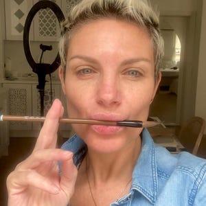 May include: A brown-handled paintbrush with a dark ferrule held horizontally between the lips. The brush features a pointed tip and a metallic blue band near the end of the handle. The handle is smooth and cylindrical in shape.
