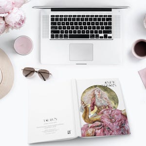 May include: A flat lay arrangement featuring a silver laptop, a beige fedora, pink peonies, a pink candle, brown sunglasses, a pink mug, two white donuts on a napkin, and an open book displaying a Taurus illustration and the text Angelic Mayers.
