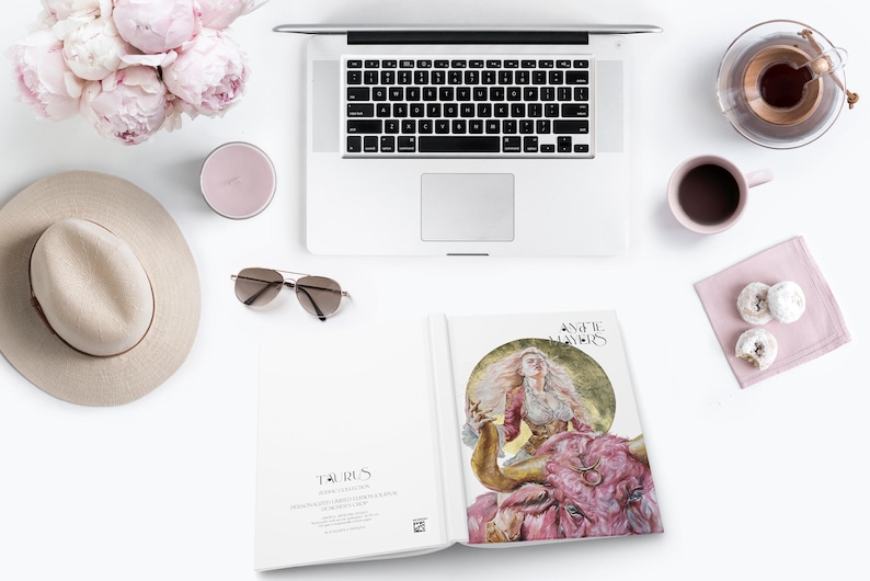 May include: A flat lay arrangement featuring a silver laptop, a beige fedora, pink peonies, a pink candle, brown sunglasses, a pink mug, two white donuts on a napkin, and an open book displaying a Taurus illustration and the text Angelic Mayers.