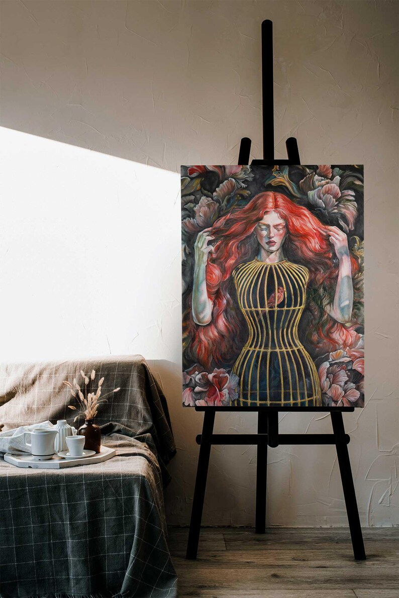 Op de afbeelding: Een schilderij op een zwarte ezel toont een persoon met lang, golvend rood haar en gesloten ogen. De persoon draagt een geel, kooivormig lijfje over het bovenlichaam, waarin een kleine vogel zichtbaar is. De achtergrond bevat donkere, weelderige bloemen.