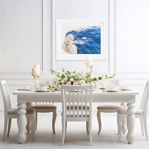 May include: A framed rectangular wall art print featuring a portrait of a person with long, flowing blue hair and closed eyes, with a small white paper boat resting in the hair, displayed in a white frame on a white paneled wall above a dining table.