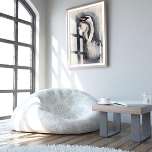 May include: A low-profile, rounded bean bag chair featuring a light gray and white patterned fabric. The chair is positioned in a room with a framed wall art piece depicting a swan and a human profile, alongside a small wooden side table.