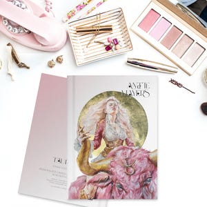 May include: A hardcover book titled 'Annette Mayers' featuring a central illustration of a person with long blonde hair riding a large pink bull against a gold circular background. The book cover includes the Taurus zodiac symbol on the bull's forehead.