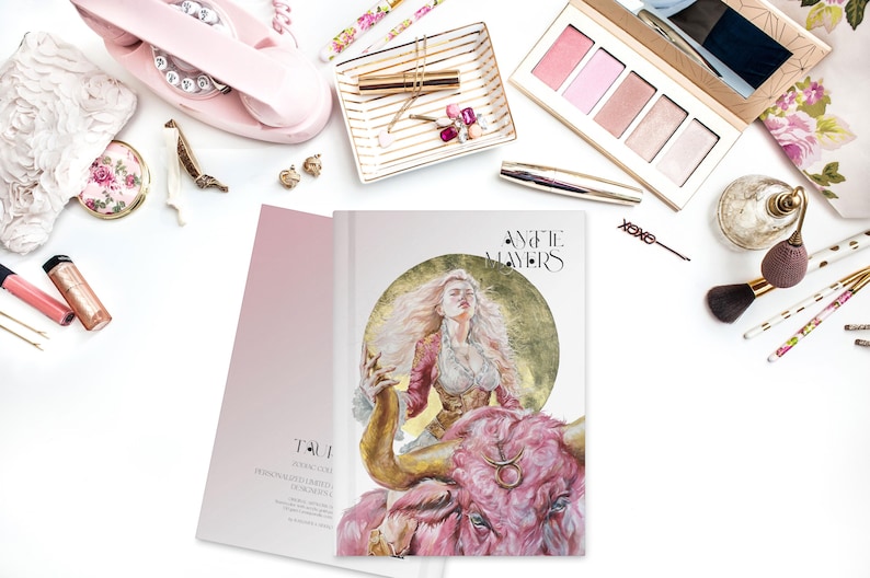 May include: A hardcover book titled 'Annette Mayers' featuring a central illustration of a person with long blonde hair riding a large pink bull against a gold circular background. The book cover includes the Taurus zodiac symbol on the bull's forehead.