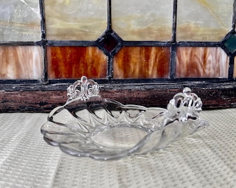 May include: A clear glass dish with a scalloped edge and ornate, decorative handles. The dish is sitting on a textured surface.