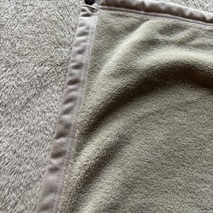 May include: A sage green towel with a soft, plush texture and a smooth, matching fabric border along the edge. A single, small, dark brown circular snap button is attached to the top corner of the border, securing the folded fabric edge.