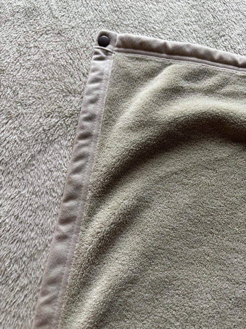 May include: A sage green towel with a soft, plush texture and a smooth, matching fabric border along the edge. A single, small, dark brown circular snap button is attached to the top corner of the border, securing the folded fabric edge.