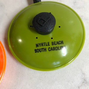 May include: Green plastic souvenir frying pan keychain with a black plastic handle and a black knob. The pan has the text "Myrtle Beach South Carolina" printed on it.