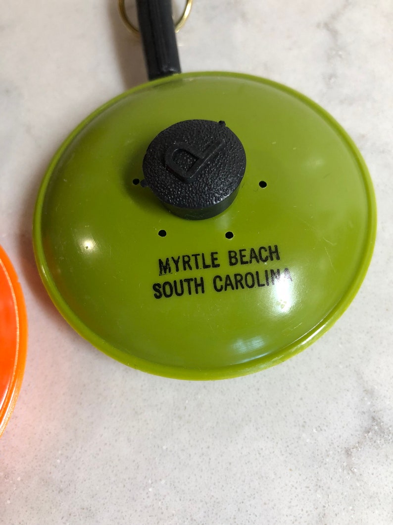 May include: Green plastic souvenir frying pan keychain with a black plastic handle and a black knob. The pan has the text "Myrtle Beach South Carolina" printed on it.
