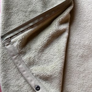 May include: A soft, light beige fleece blanket featuring a folded corner that reveals a matching fabric trim. The edge is secured with a long, integrated zipper and a single dark-colored metal snap button, providing a functional closure mechanism.