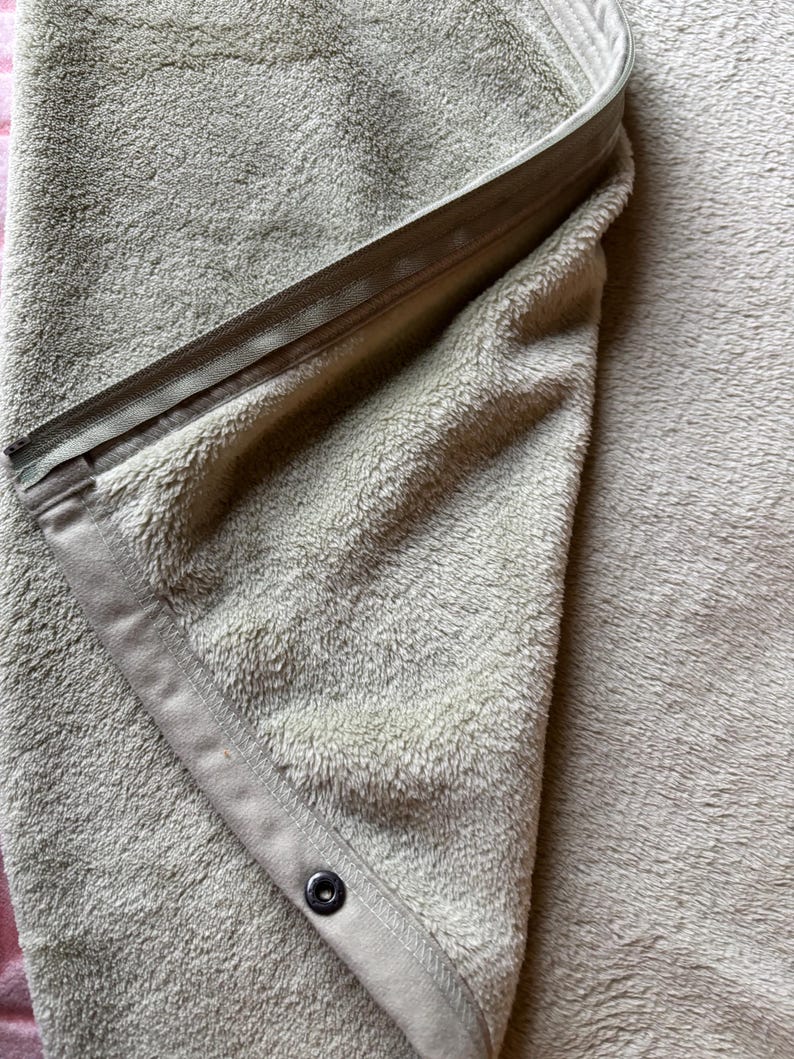 May include: A soft, light beige fleece blanket featuring a folded corner that reveals a matching fabric trim. The edge is secured with a long, integrated zipper and a single dark-colored metal snap button, providing a functional closure mechanism.