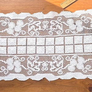May include: White lace table runner with a floral and geometric design. The runner features a rectangular center with a grid pattern, framed by intricate floral and scrollwork patterns along the edges. The runner is displayed on a wooden surface.