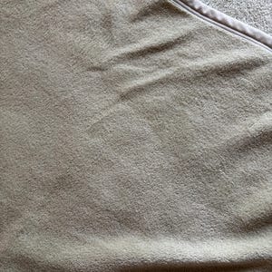 May include: A soft, light brown blanket with a plush, fuzzy texture and a smooth, finished edge. The fabric features a dense, short-pile surface that creates subtle shadows and highlights across the folded material, showing a uniform color.