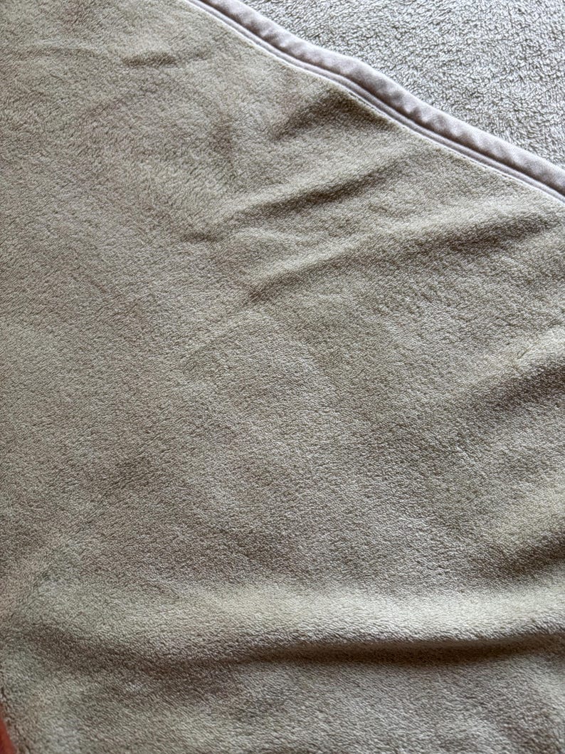 May include: A soft, light brown blanket with a plush, fuzzy texture and a smooth, finished edge. The fabric features a dense, short-pile surface that creates subtle shadows and highlights across the folded material, showing a uniform color.