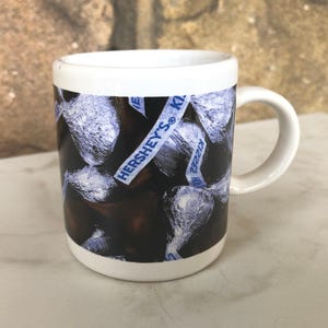 May include: White ceramic mug with a black and white image of Hershey's Kisses wrapped in silver foil. The mug has a blue and white label that says "Hershey's Kisses".