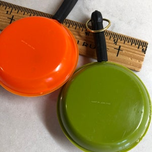 May include: Two miniature toy frying pans with black handles and gold key rings. One pan is orange and the other is green. The bottom of each pan has a label with the text "Made in USA" and a number.