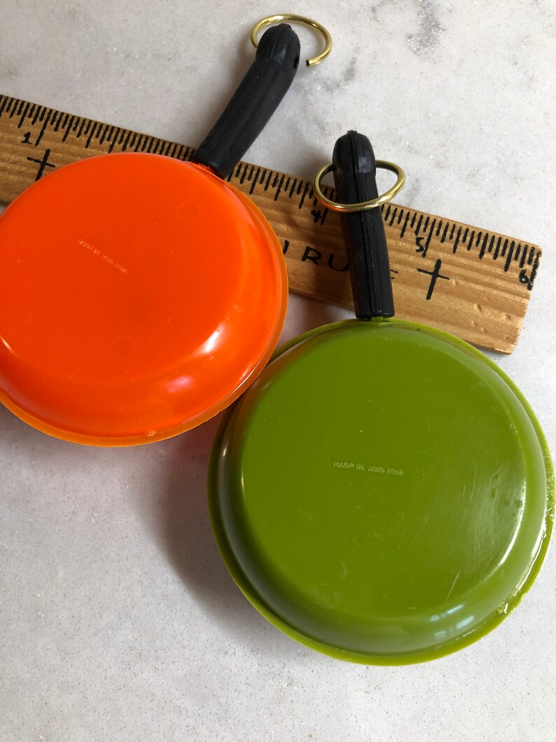May include: Two miniature toy frying pans with black handles and gold key rings. One pan is orange and the other is green. The bottom of each pan has a label with the text "Made in USA" and a number.