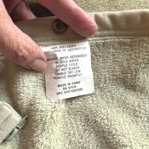 May include: A beige garment with a soft, fuzzy interior lining and a metal snap button. A white care label is attached, reading: 100% POLYESTER, EXCLUSIVE OF DECORATION, MACHINE WASH SEPERATELY IN COLD WATER, GENTLE CYCLE, DO NOT BLEACH, TUMBLE DRY LOW, REMOVE PROMPTLY, MADE IN CHINA, RN 91519, UPC: 675716231552.