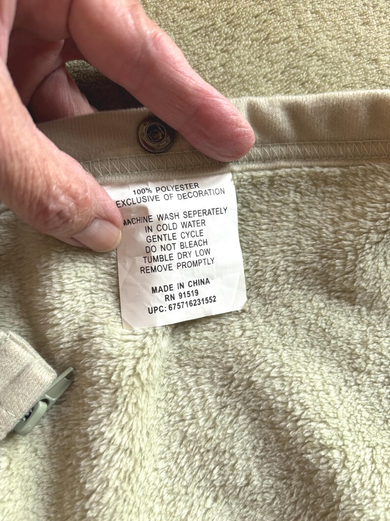May include: A beige garment with a soft, fuzzy interior lining and a metal snap button. A white care label is attached, reading: 100% POLYESTER, EXCLUSIVE OF DECORATION, MACHINE WASH SEPERATELY IN COLD WATER, GENTLE CYCLE, DO NOT BLEACH, TUMBLE DRY LOW, REMOVE PROMPTLY, MADE IN CHINA, RN 91519, UPC: 675716231552.