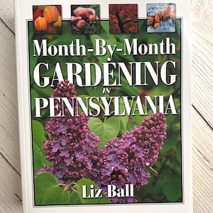 May include: A hardcover book titled Month-By-Month Gardening in Pennsylvania by Liz Ball. The cover features a large cluster of purple lilac flowers with green leaves, topped by four small square photographs of seasonal garden scenes.