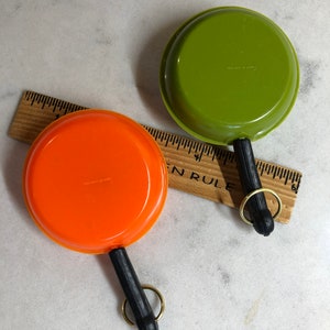 May include: Two miniature toy frying pans with black handles and gold key rings. One pan is orange and the other is green.