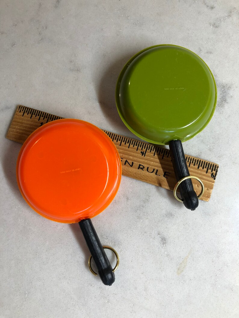May include: Two miniature toy frying pans with black handles and gold key rings. One pan is orange and the other is green.