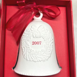 May include: A white ceramic bell ornament featuring a raised relief wreath design and the red text 2007 in the center. The bell is topped with a red satin ribbon bow and is presented inside a red box with a matching red interior lining.