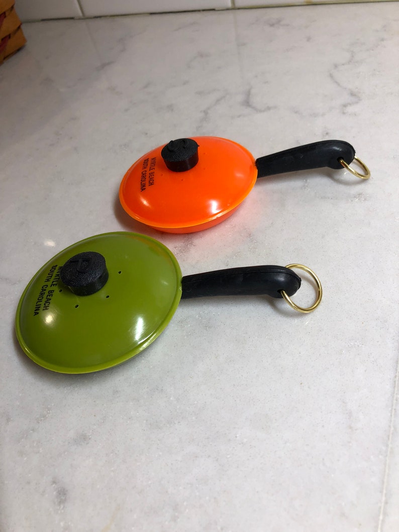 May include: Two small orange and green plastic frying pan keychains with black handles and gold rings. The orange pan has the text "Myrtle Beach South Carolina" printed on it. The green pan has the text "Myrtle Beach South Carolina" printed on it.