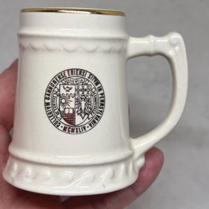 May include: A cream-colored ceramic beer stein featuring a gold-trimmed rim and a handle. The front displays a circular emblem with the text COLLEGIUM CANNONENSE ERIENSI SITUM IN PENNSYLVANIA MCMXLIV and a central shield with various symbols.