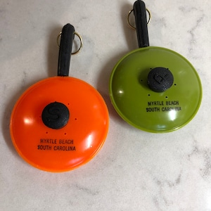 May include: Two small, plastic frying pan keychains. One is orange and the other is green. Both have black handles and say "Myrtle Beach South Carolina" on the front.