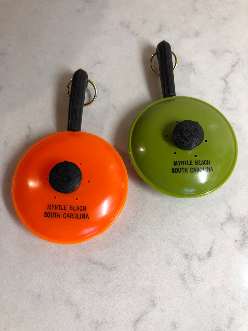 May include: Two small, plastic frying pan keychains. One is orange and the other is green. Both have black handles and say "Myrtle Beach South Carolina" on the front.