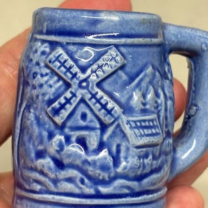 May include: Small blue ceramic mug with a handle. The mug features a raised relief design of a windmill, house, and landscape. The mug is a solid blue color with a glossy finish.