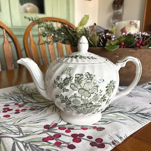 May include: White ceramic teapot with a lid, featuring a green floral pattern. The teapot has a curved spout and handle. It sits on a table runner with a red berry and green leaf design. The background includes a wooden bowl and a green cabinet.