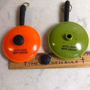 May include: Two small orange and green plastic frying pans with black handles and a black knob on the top. The pans have the text "Myrtle Beach South Carolina" on the bottom.