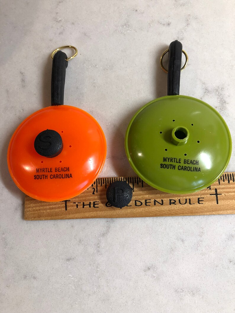 May include: Two small orange and green plastic frying pans with black handles and a black knob on the top. The pans have the text "Myrtle Beach South Carolina" on the bottom.