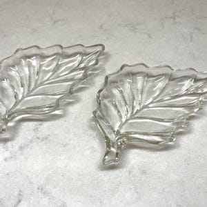 May include: Two clear glass leaf-shaped dishes. The dishes have detailed veining and a scalloped edge, giving them a realistic appearance. They are placed on a light-colored, textured surface.