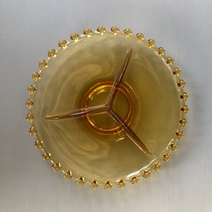 May include: Amber-colored glass serving dish with three sections, featuring a central ring and three dividers. The rim is adorned with small, rounded decorative elements. The dish is designed for serving snacks or appetizers.