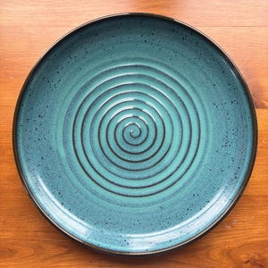 May include: A round, teal-colored ceramic plate with a spiral design in the center. The plate has a dark brown rim and a speckled texture. The plate is on a wooden surface.