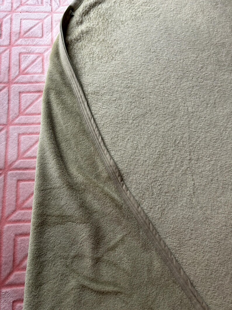 May include: A sage green blanket with a soft, plush, fuzzy texture. The blanket features a smooth, matching sage green fabric trim along the curved edge, providing a clean finish to the overall design of this cozy, solid-colored textile item.
