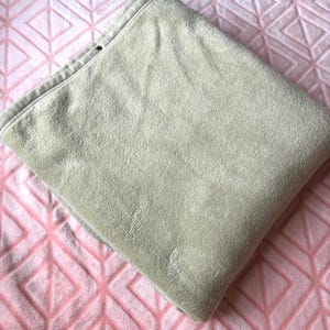 May include: A folded, light beige, plush blanket with a soft, velvety texture. The blanket features a finished hem along the top edge with a small, dark circular detail visible near the corner, showcasing a smooth, consistent fabric surface.