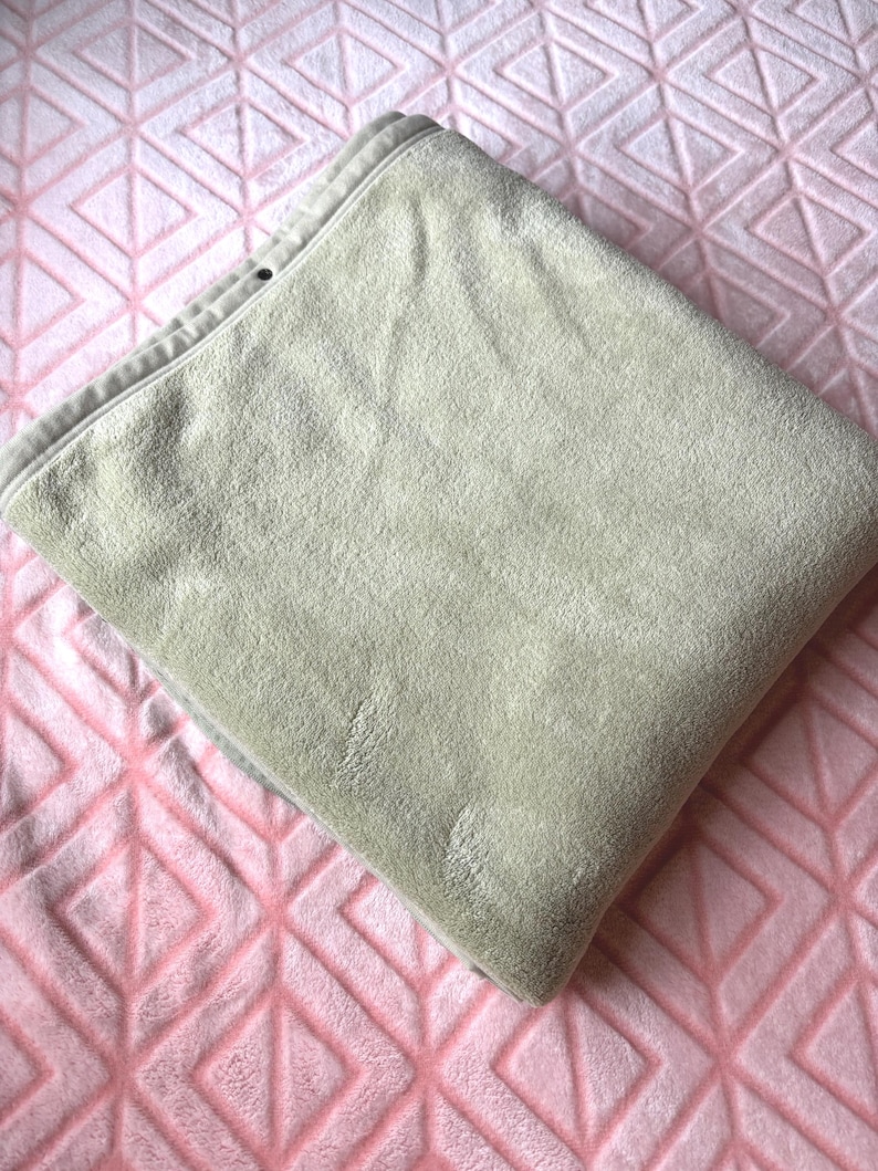 May include: A folded, light beige, plush blanket with a soft, velvety texture. The blanket features a finished hem along the top edge with a small, dark circular detail visible near the corner, showcasing a smooth, consistent fabric surface.