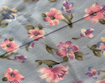 May include: Close-up of a light blue fabric with a floral pattern. The flowers are in shades of pink, purple, and blue, with green leaves. The fabric has a subtle, textured appearance, creating a vintage aesthetic.