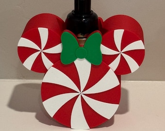 May include: A red and white peppermint-themed soap dispenser with a black pump. The dispenser is decorated with a green bow and three red and white peppermint candy shapes, resembling Mickey Mouse ears. The design is festive and suitable for the holiday season.