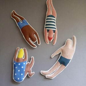 May include: Four fabric figures in various diving poses. One figure is brown with blue shorts, another has a blue and white striped swimsuit, a third has a blue polka dot swimsuit, and the last is a light skin tone with blue shorts. All figures have white stitching.