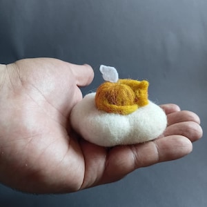 May include: A needle-felted sculpture of a small orange cat curled up asleep on a round, fluffy white cloud-like base. The cat has a tiny white wing protruding from its back, and the entire piece is crafted from soft, textured wool fibers.