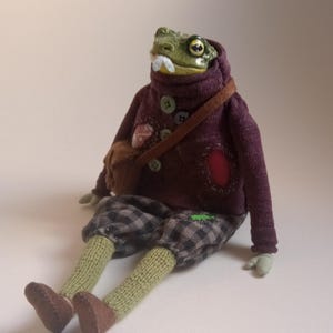 May include: A whimsical frog doll with a green head, a white mustache, and a burgundy sweater. The doll wears gray plaid pants, green socks, and brown shoes. It has a brown satchel bag and is sitting down.
